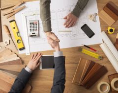 Architect and customer businessman shaking hands top view, desktop with building project, tools and wood swatches on background